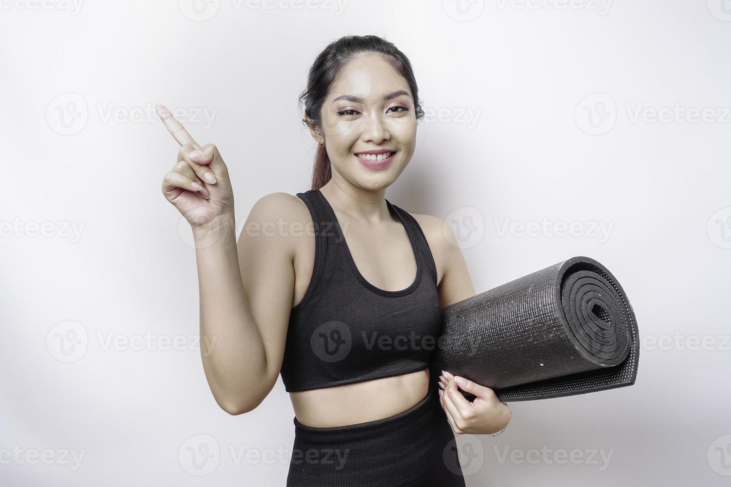 upphetsad sportig asiatisk kvinna bär sportkläder pekande på de kopia Plats på topp av henne medan bärande yoga matta, isolerat förbi vit bakgrund foto