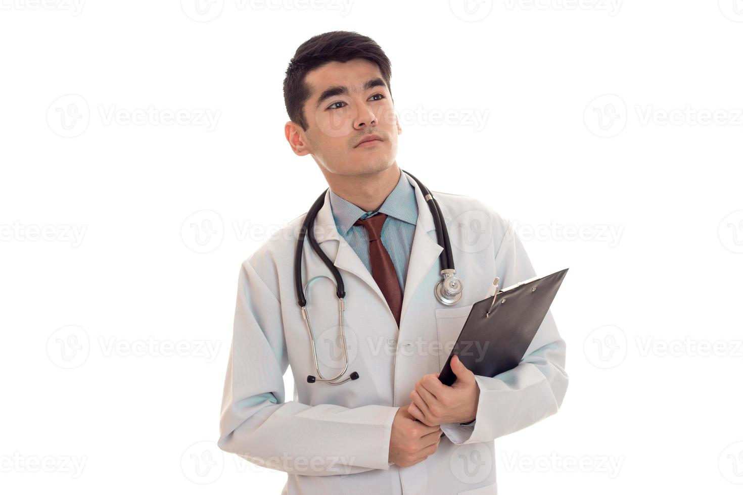 ung skön brunett man läkare i enhetlig med stetoskop på hans axlar göra anteckningar isolerat på vit bakgrund foto