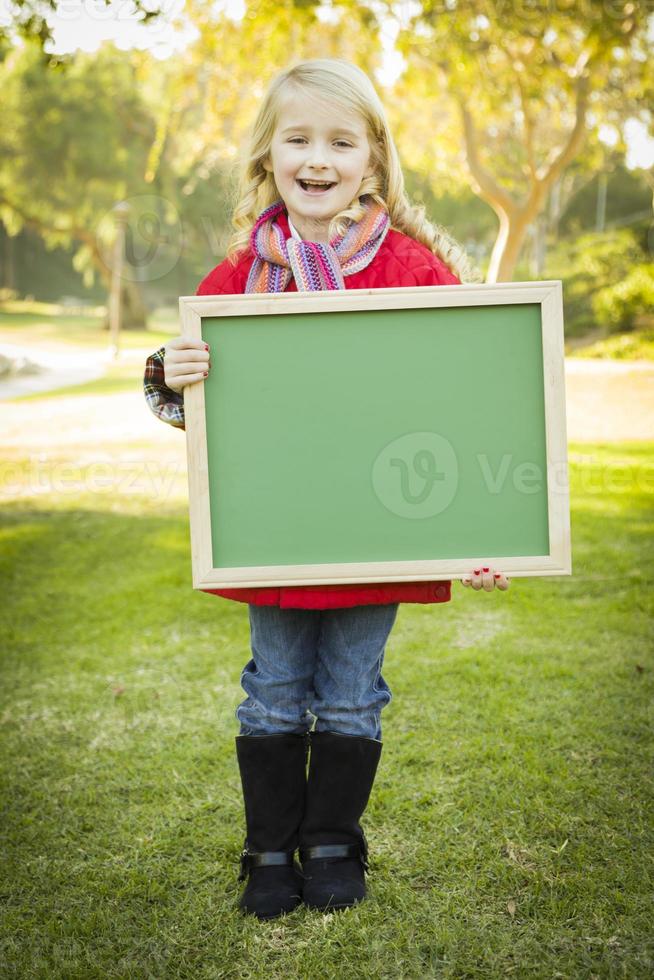 söt flicka innehav en grön svarta tavlan bär vinter- täcka utomhus foto