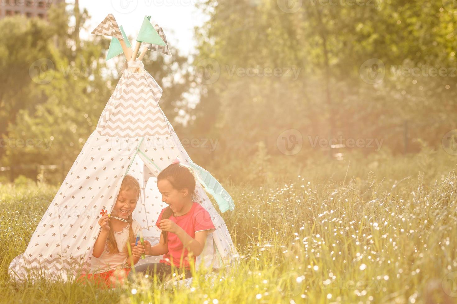 två Lycklig skrattande liten flickor i camping tält i maskros fält foto