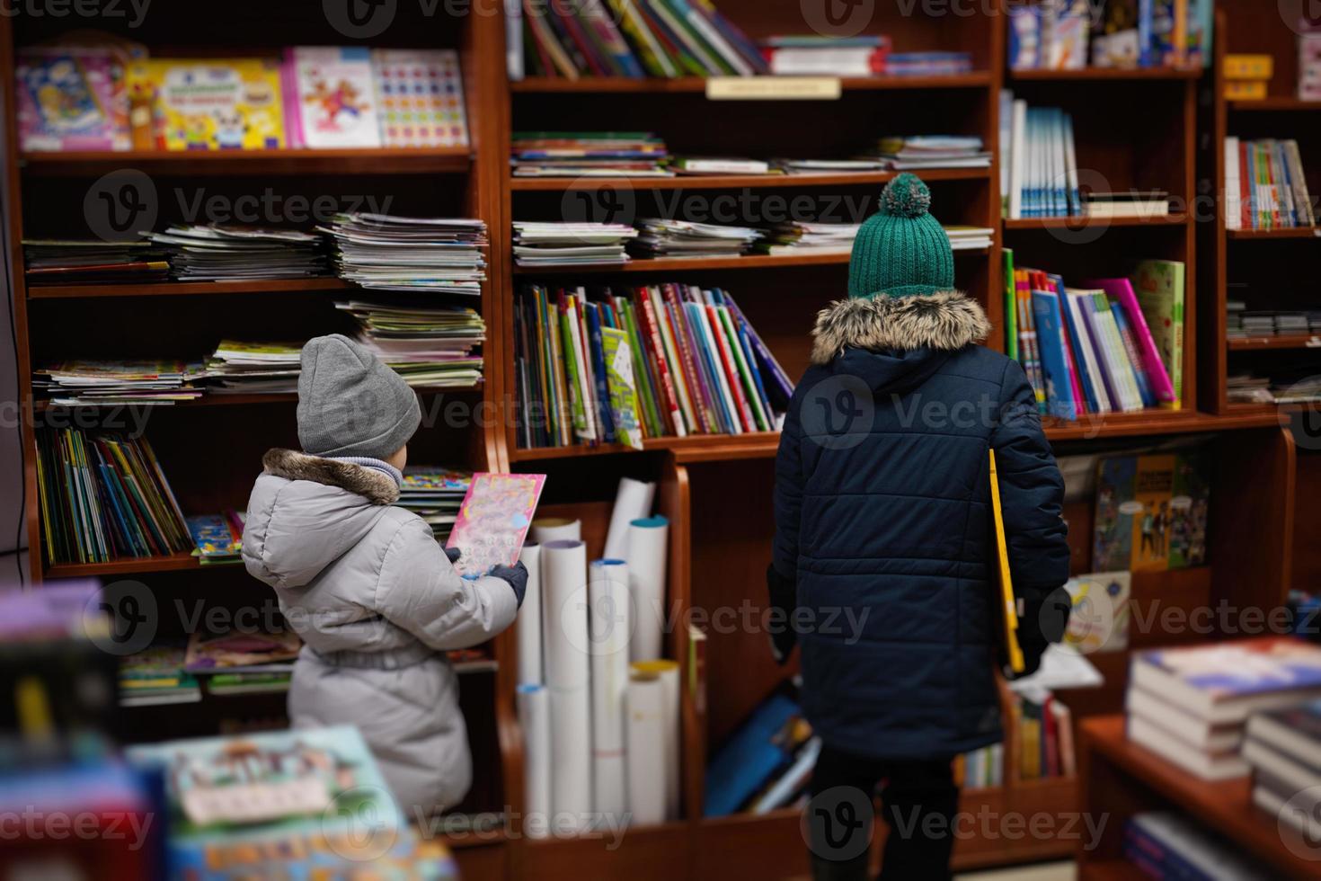 liten syster med bror i jacka nå en bok från bokhylla på de bibliotek. inlärning och utbildning av europeisk ungar. foto