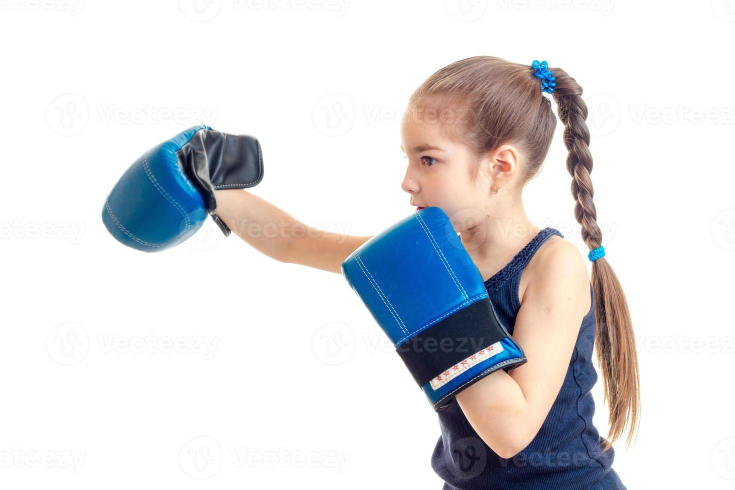 liten flicka med en hårpiska står sidled i främre av de kamera och sträckt framåt- hand i stor boxning handskar foto