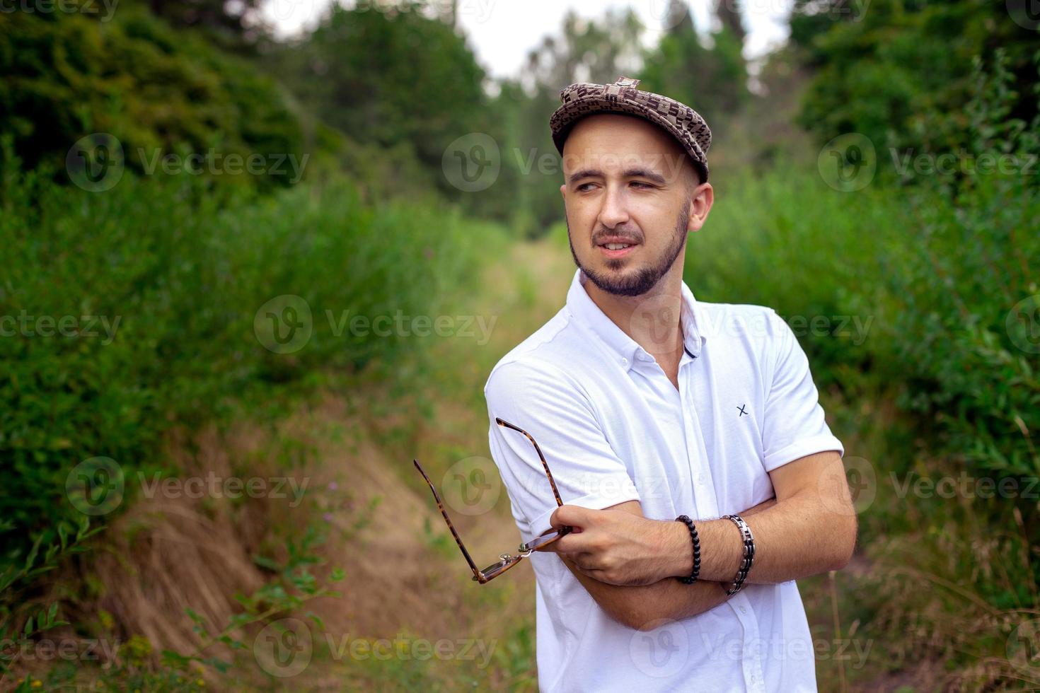 sexig man modell i hatt innehar solglasögon och ser bort på de grön trädgård foto