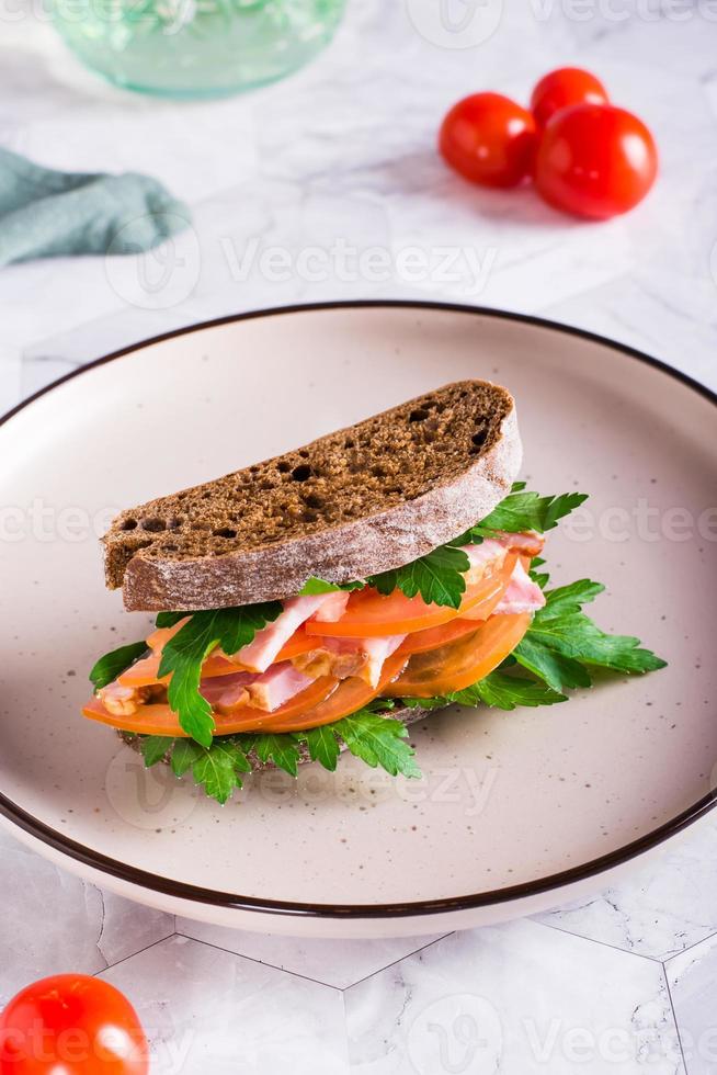 hemlagad skinka, tomat och grönt smörgås på råg rostat bröd på en tallrik på de tabell. vertikal se foto