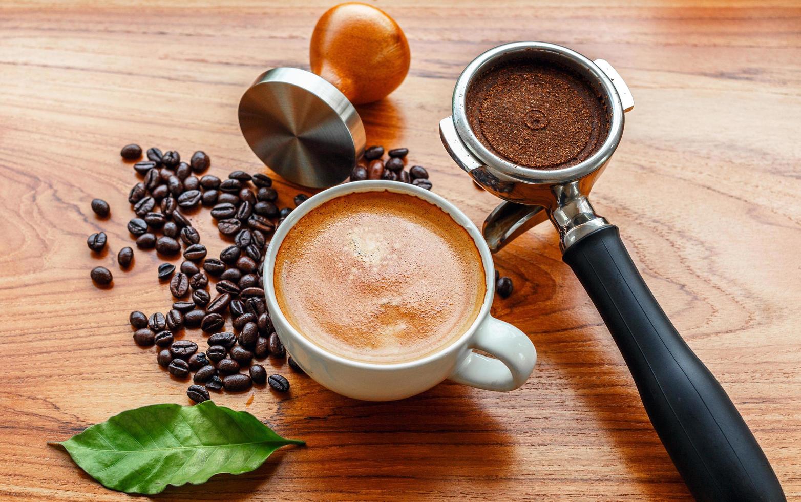 topp se av espresso kaffe kopp och Utrustning av Barista kaffe verktyg portafilter med manipulera och mörk rostad kaffe bönor med grön kaffe blad på trä- tabell foto