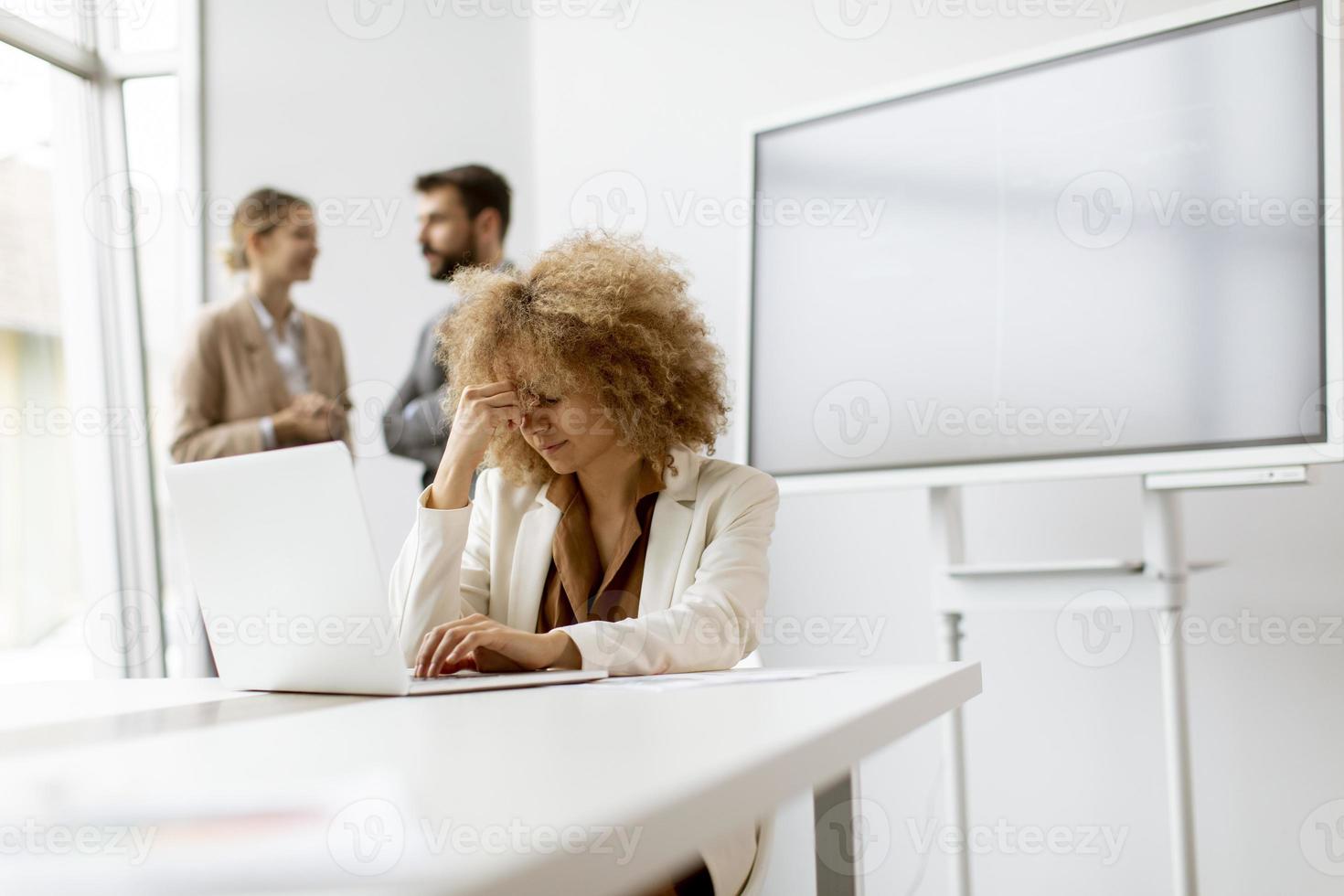 ung worrie affärskvinna använder sig av bärbar dator i de kontor med ung människor Arbetar Bakom henne foto