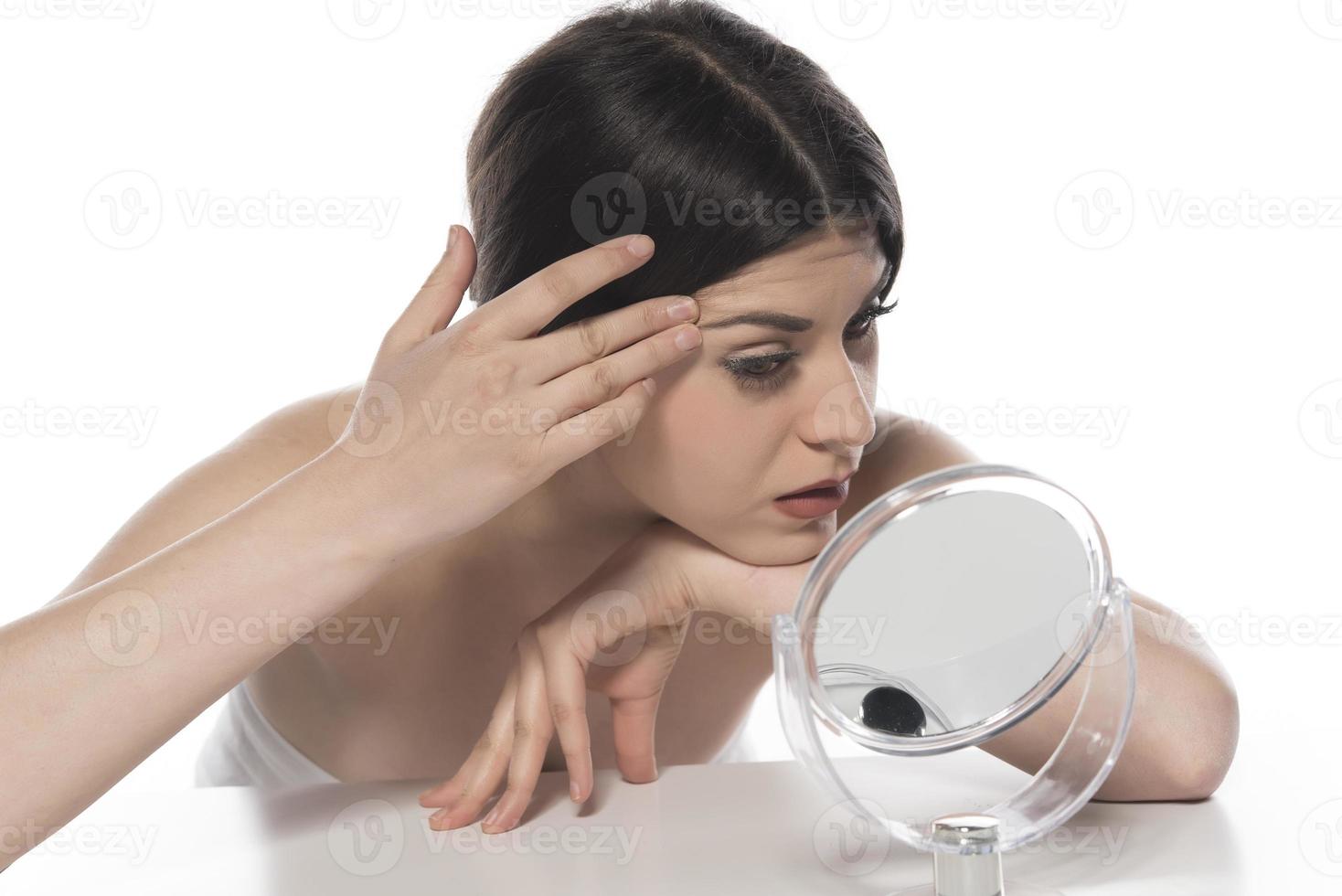 skön attraktiv brunett kvinna kontroll henne ansikte hud i främre av en spegel på vit bakgrund foto
