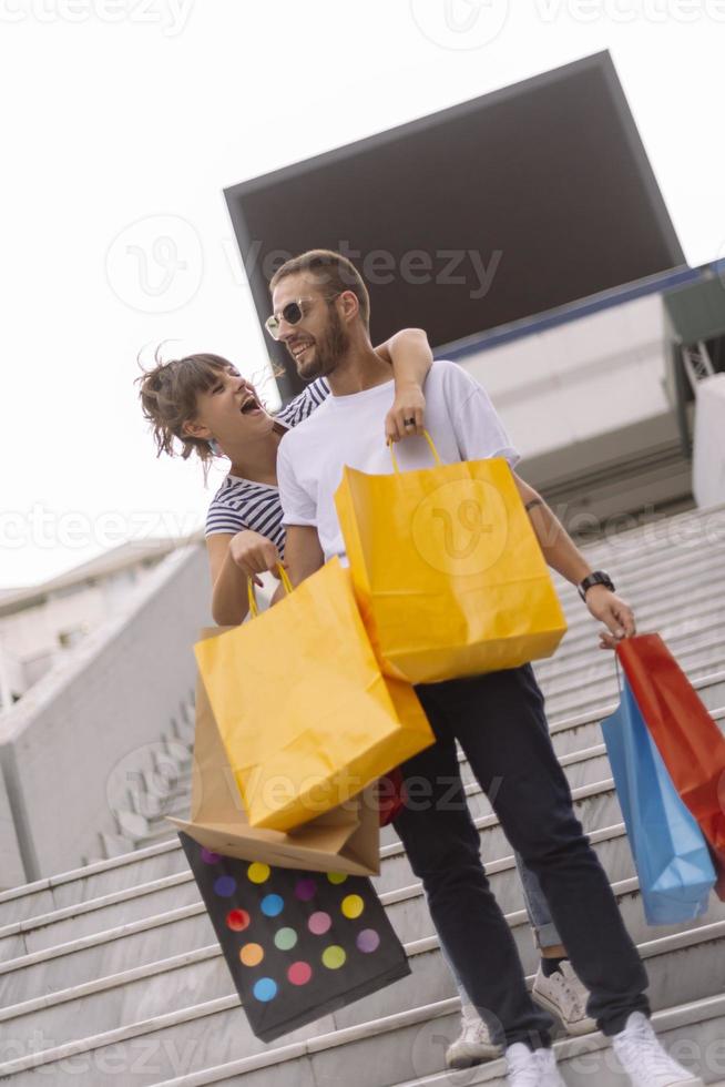 porträtt av glad caucasian ung par man och kvinna innehav många papper påsar efter handla medan gående och talande på gata. Lycklig familj par med paket utomhus. uppköp begrepp foto