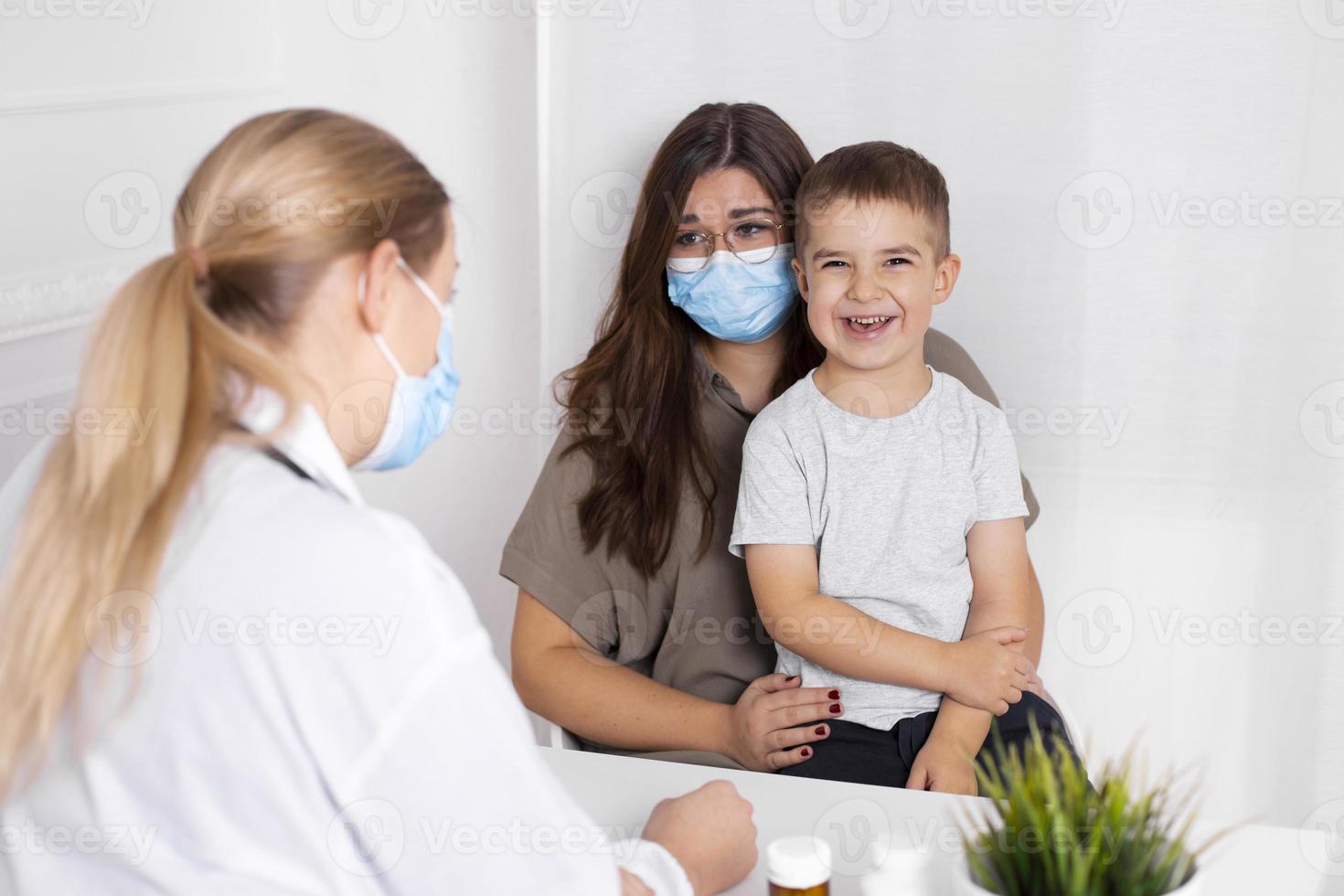 ung brunett kvinna med liten pojke har samråd på barnläkare kontor. läkare, barn och mor bär ansiktsmasker under coronavirus och influensa utbrott. virus skydd. covid 19. foto