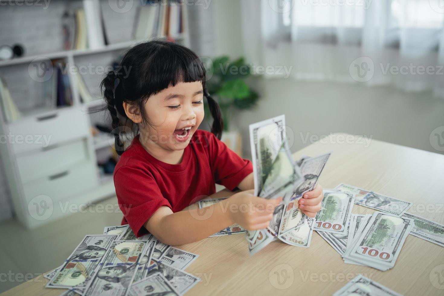 asiatisk bebis flicka bär en röd t-shirt innehav dollar räkningen på trä tabell skrivbord i levande rum på Hem. sparande investering rikedom, tillväxt stock, investera intressera, fond, sparande pengar för de framtida begrepp. foto