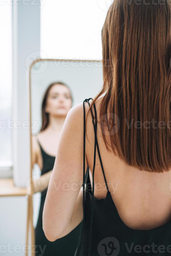 ung kvinna med lång mörk hår på kväll elegant svart klänning med öppen tillbaka nära spegel i de vit interiör, människor från Bakom foto