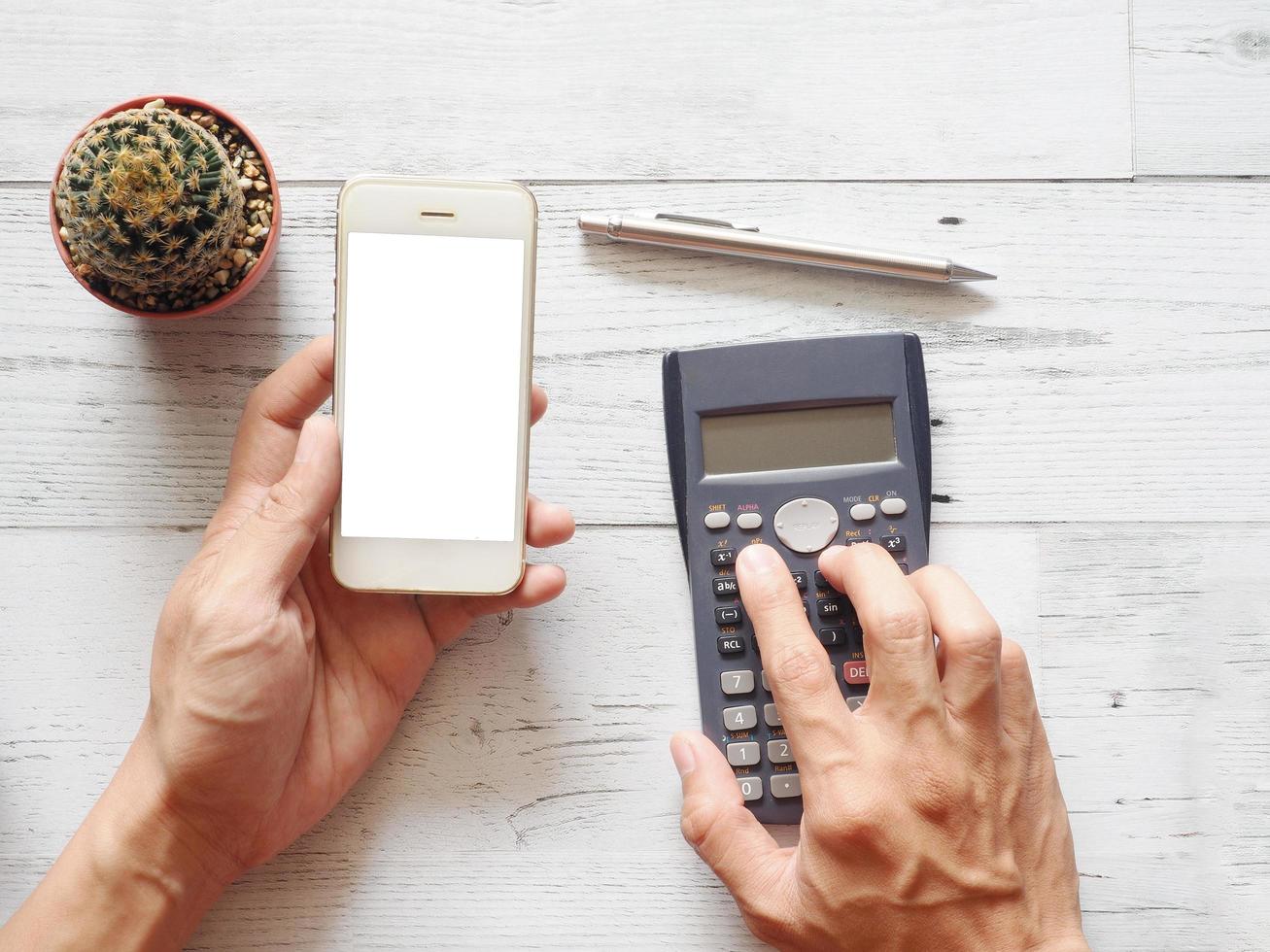 hand innehav mobiltelefon vit skärm och använder sig av kalkylator med kaktus och silver- penna på vit trä tabell natur skugga och solljus topp se och Plats företag begrepp foto