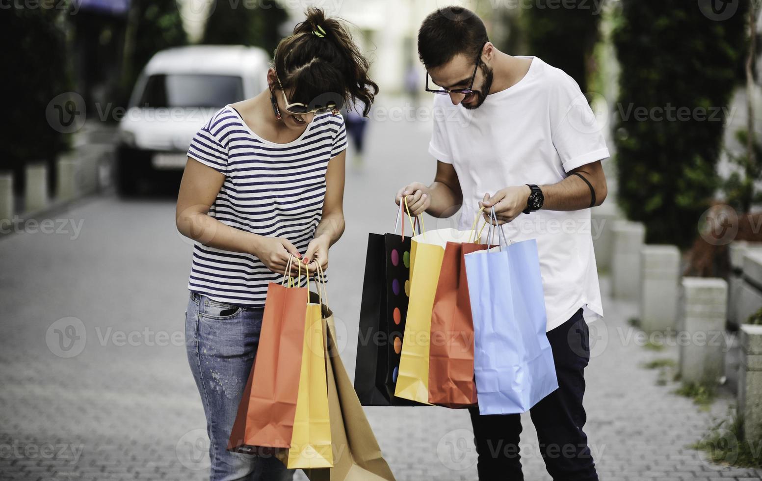 porträtt av glad caucasian ung par man och kvinna innehav många papper påsar efter handla medan gående och talande på gata. Lycklig familj par med paket utomhus. uppköp begrepp foto