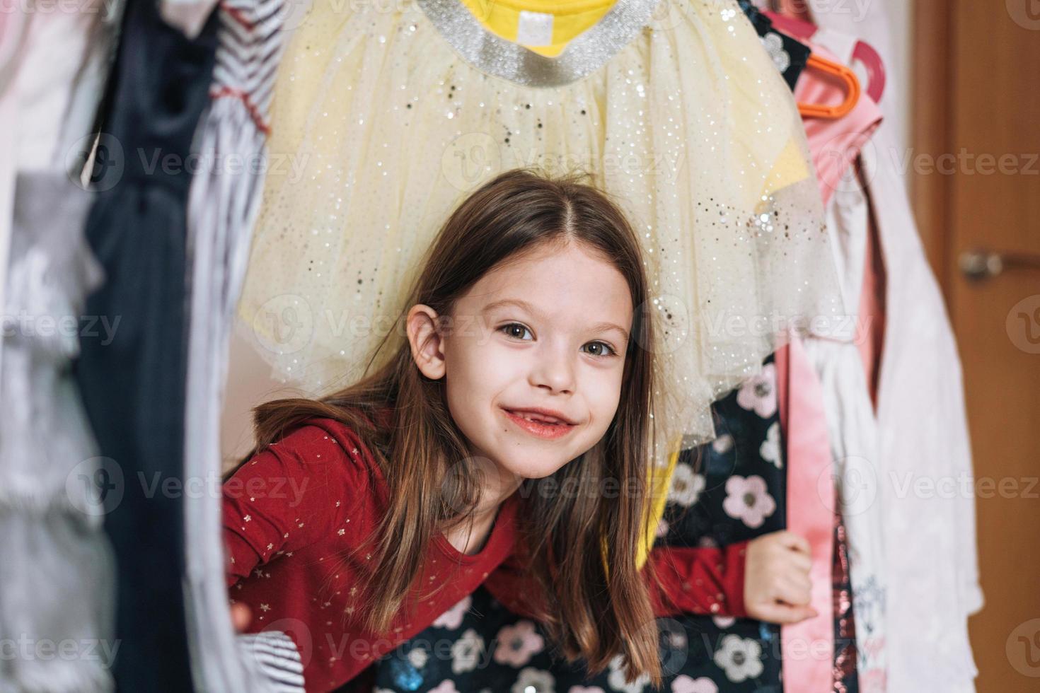 leende mellan liten flicka med lång mörk hår i röd klänning bland henne skön klänningar i garderob i de barns rum på Hem foto