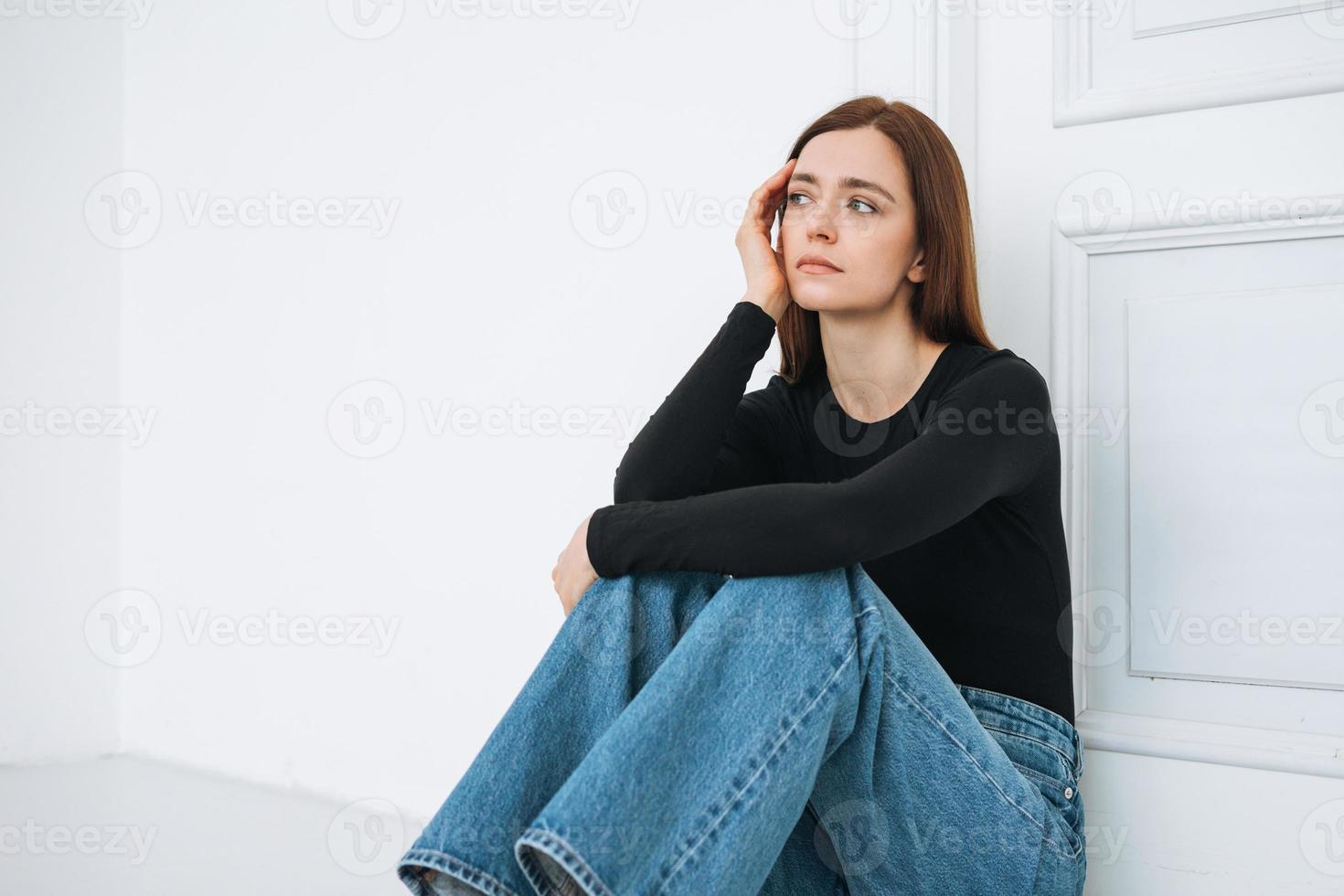 ung olycklig kvinna tonåring flicka med lång hår i jeans Sammanträde på golv i stängd placera förbi dörr på Hem, negativ känsla foto