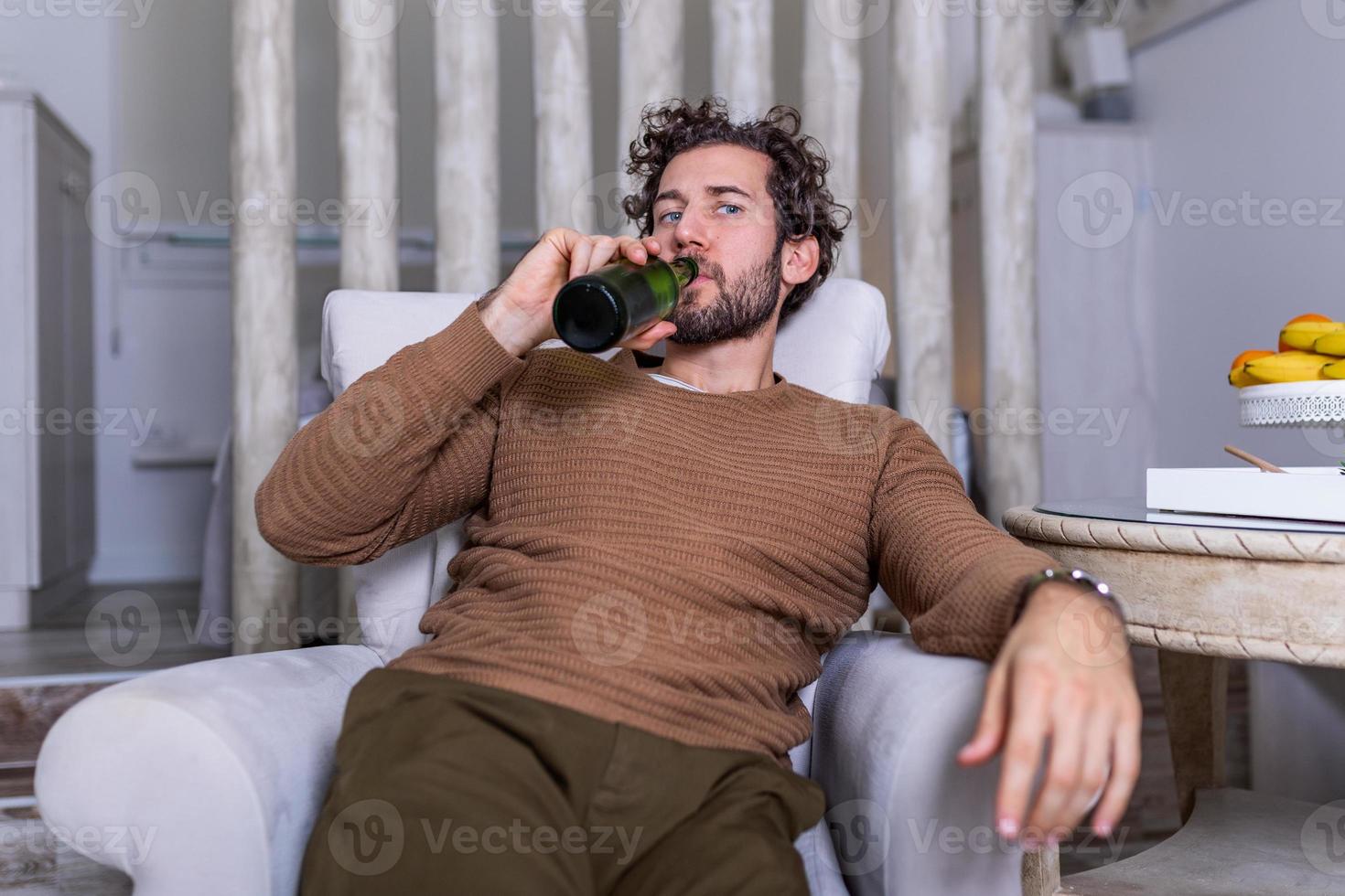 efter arbete en deprimerad kille Sammanträde på soffa, dricka en kall öl. hand innehav en flaska, man dricka Hem ensam. alkoholism, alkohol missbruk och människor begrepp foto