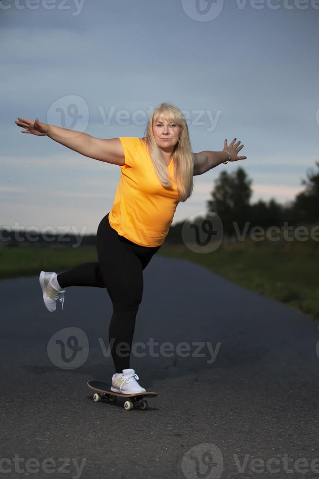 knubbig medelålders europeisk kvinna, övervikt, i en träningsoverall, lär till rida en skateboard, går i för sporter. foto