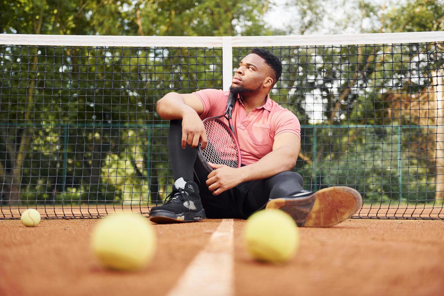 sitter nära netto och tar en ha sönder. afrikansk amerikan man i rosa skjorta sitter med tennis racket på de domstol utomhus foto