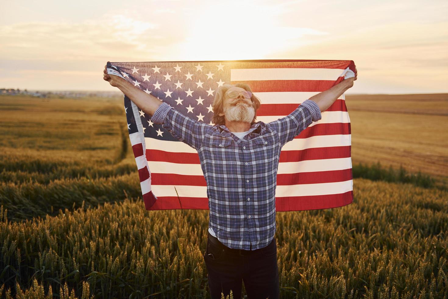 innehav USA flagga i händer. patriotisk senior eleganta man med grå hår och skägg på de jordbruks fält foto