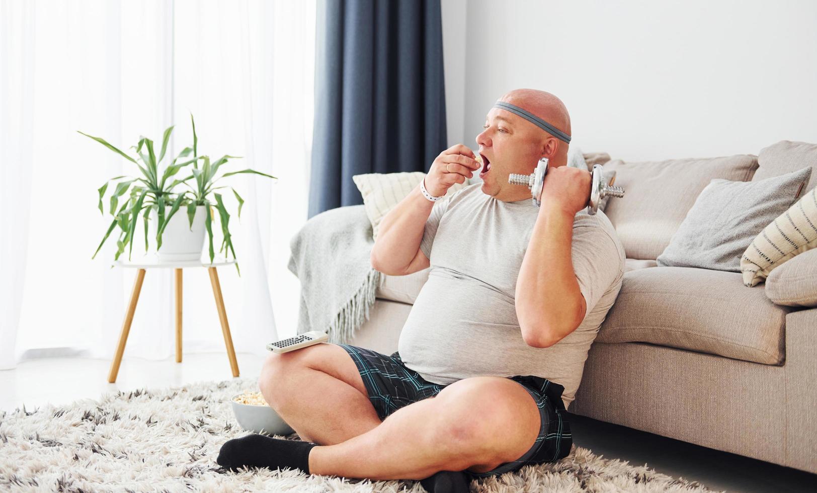 äter och gör övningar. rolig övervikt man i tillfällig kläder är inomhus på Hem foto