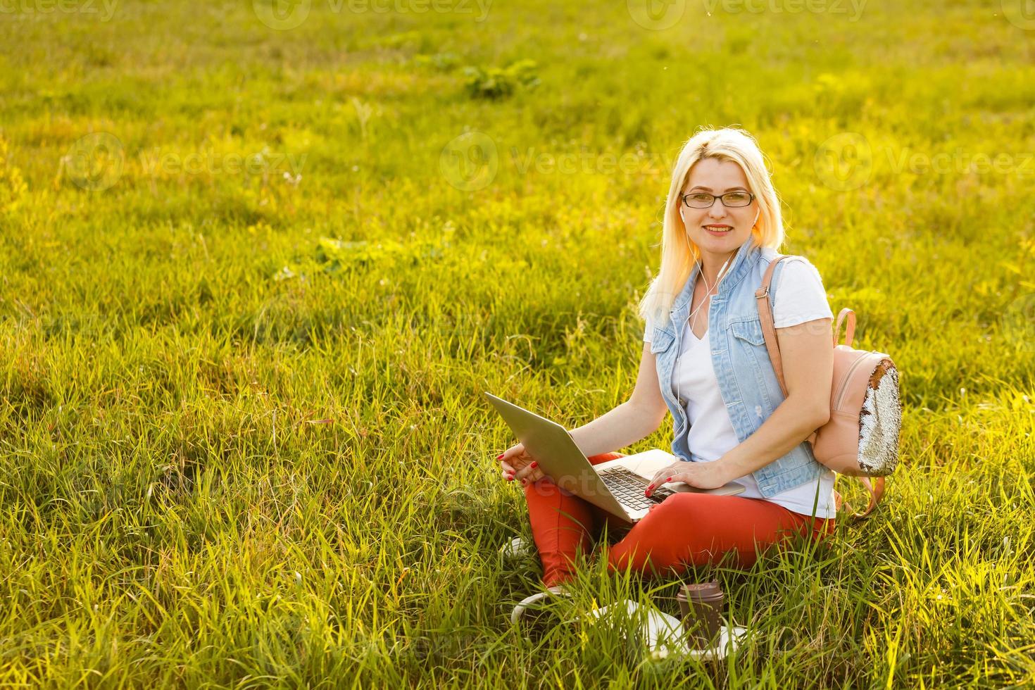 ung kvinna med bärbar dator sitter på de gräs i de parkera på en solig dag. skrattande blond i en vit tank topp och shorts. uppkopplad Träning, avlägsen arbete och kommunikation i social nät foto