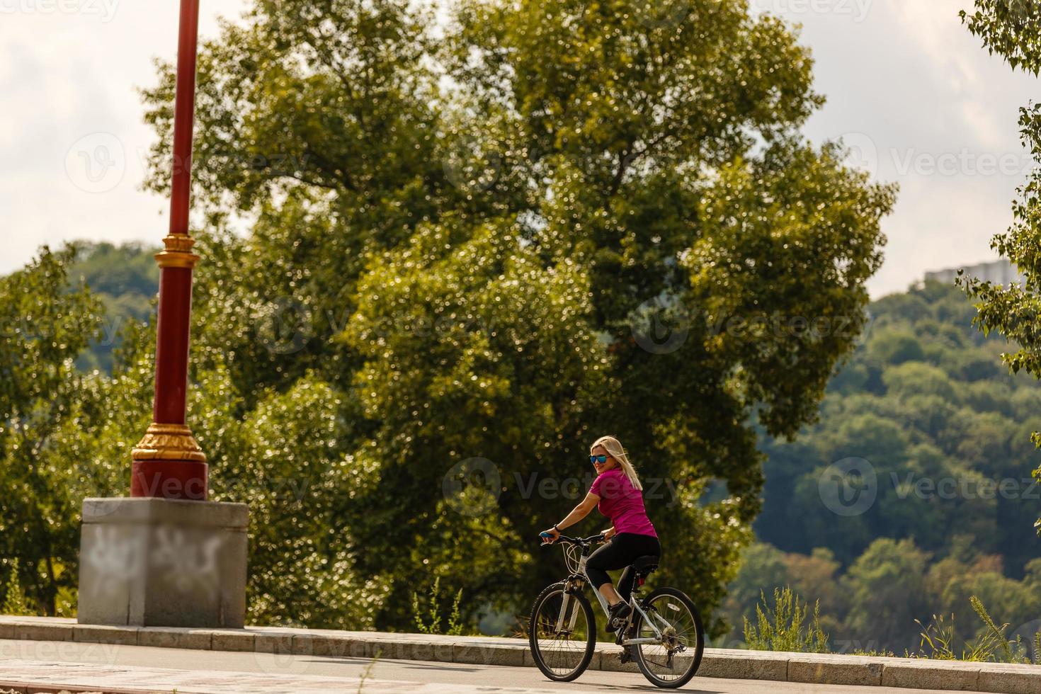 skön flicka med blond hår lyckligt ridning cykel. porträtt av ung lady i j solglasögon har roligt, ridning på cykel längs stad gator foto