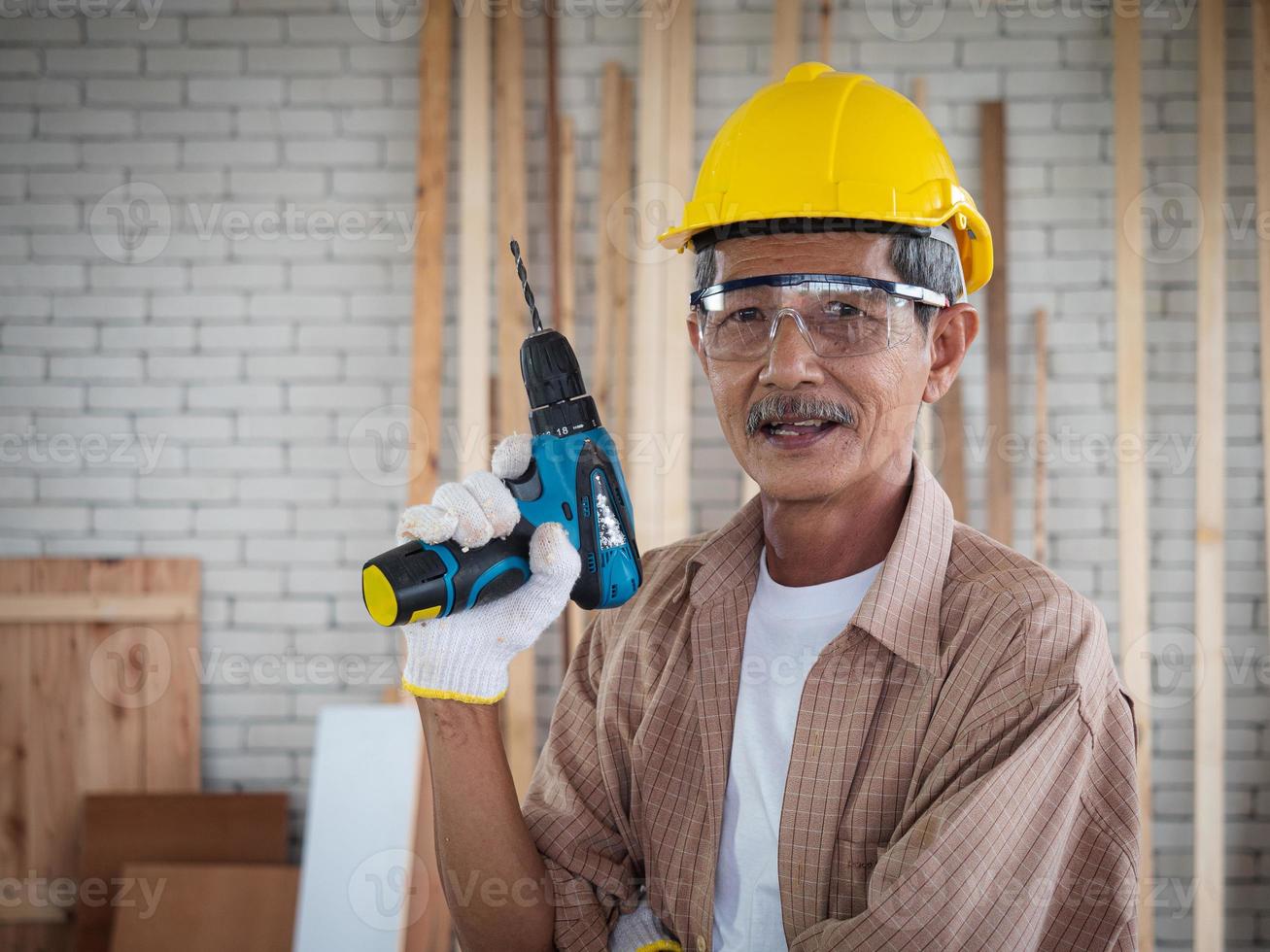porträtt av en senior snickare visa en kraft skruvmejsel till kör skruva nagel i en trä verkstad, gammal man undersöker kraft skruvmejslar, en konstruktion industri tema foto