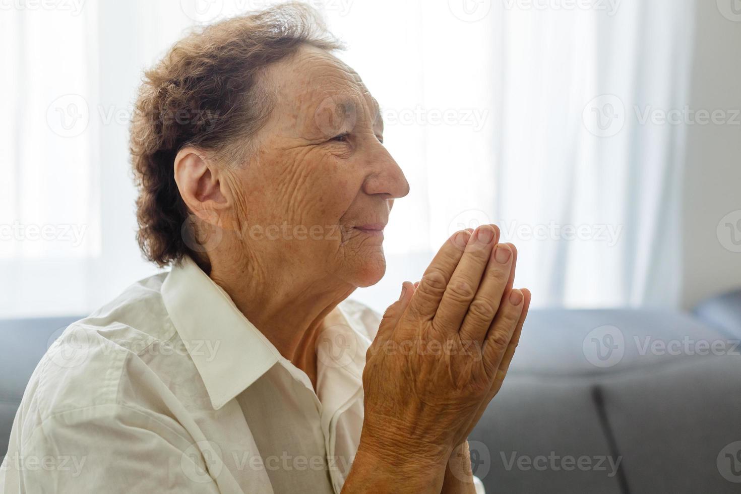 trogen gammal senior mormor be med hoppas tro innehav händer tillsammans sitta på soffa ensam på Hem, religiös äldre kvinna troende kristen med ögon stängd säga tacksamhet dyrkan bön begrepp foto
