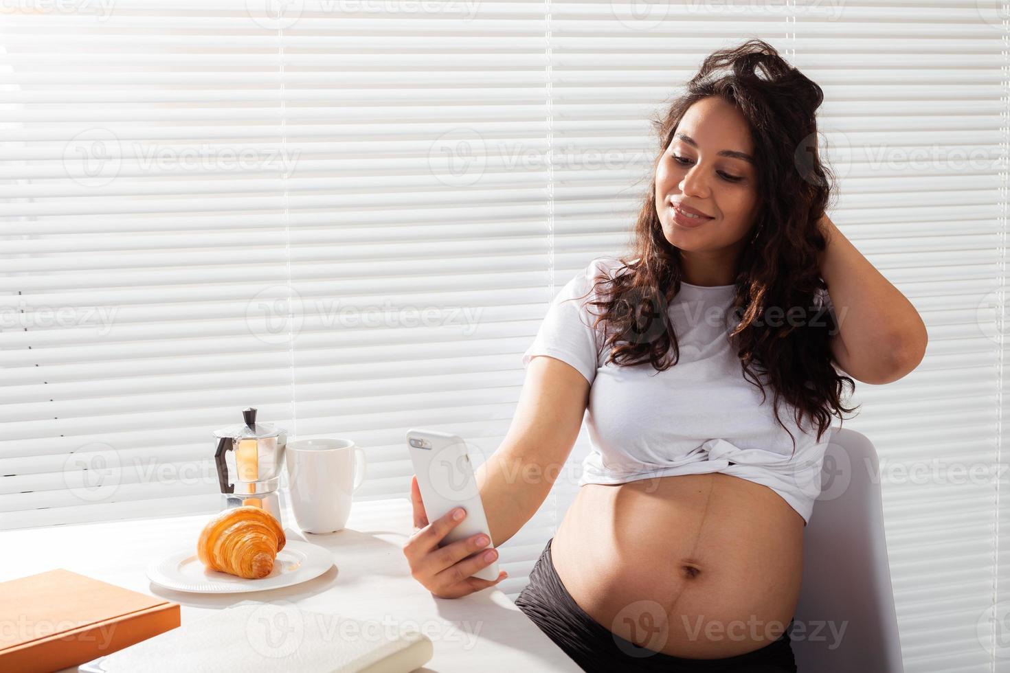 gravid kvinna tar selfie på henne smartphone medan frukost. moderskap lämna och teknik begrepp. foto