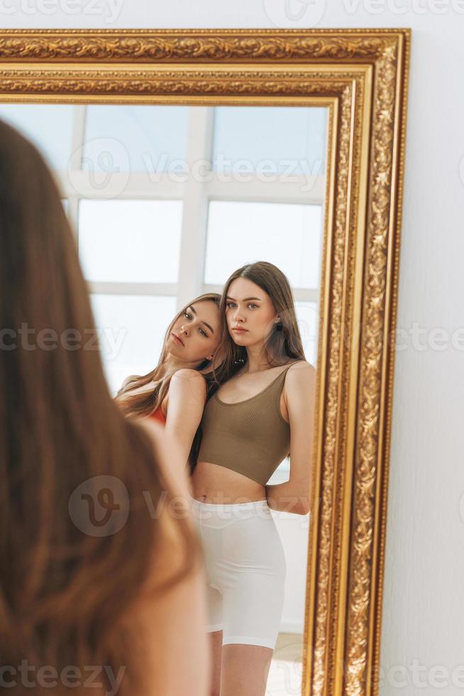 mode skönhet modeller två systrar tvillingar skön flickor med lång hår i sport ha på sig ser till spegel i de ljus studio foto