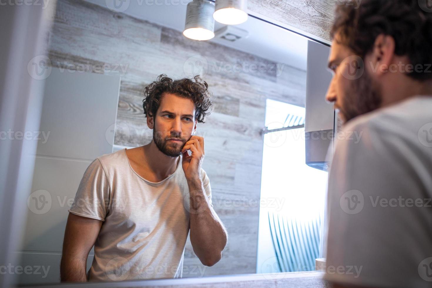 morgon- hygien, stilig man i de badrum ser i spegel. reflexion av stilig man med skägg ser på spegel och rörande ansikte i badrum grooming foto