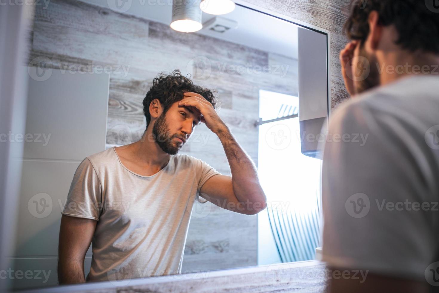 stilig ung man rörande hans hår med hand och grooming i badrum på Hem. vit metrosexual man orolig för hår förlust och ser på spegel hans avtagande hårfäste. foto