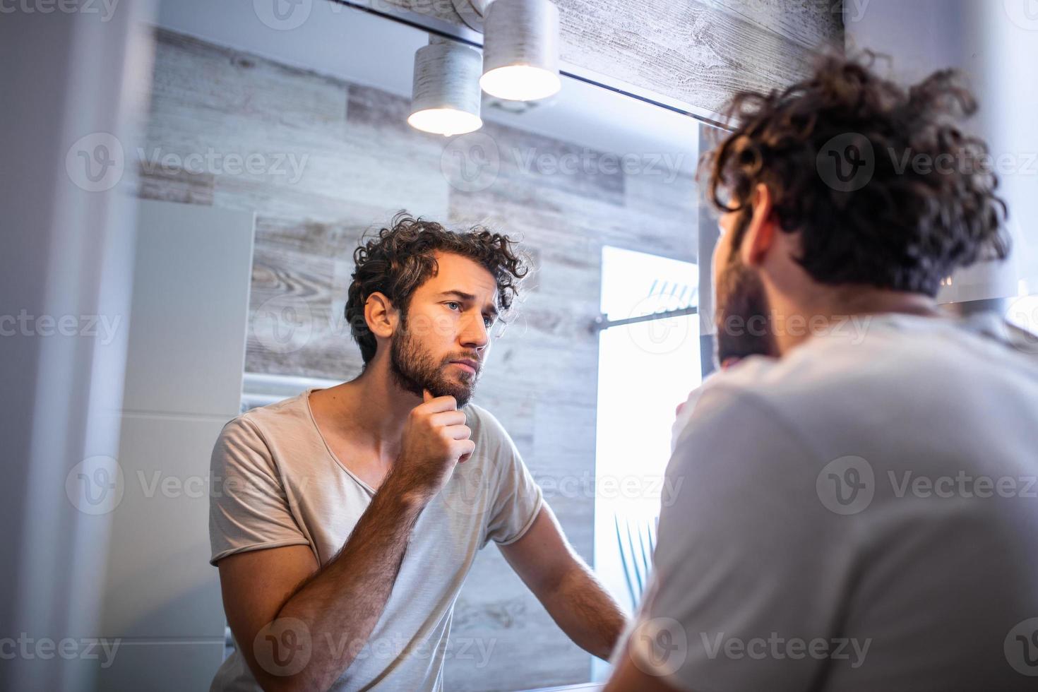 morgon- hygien, stilig man i de badrum ser i spegel. reflexion av stilig man med skägg ser på spegel och rörande ansikte i badrum grooming foto