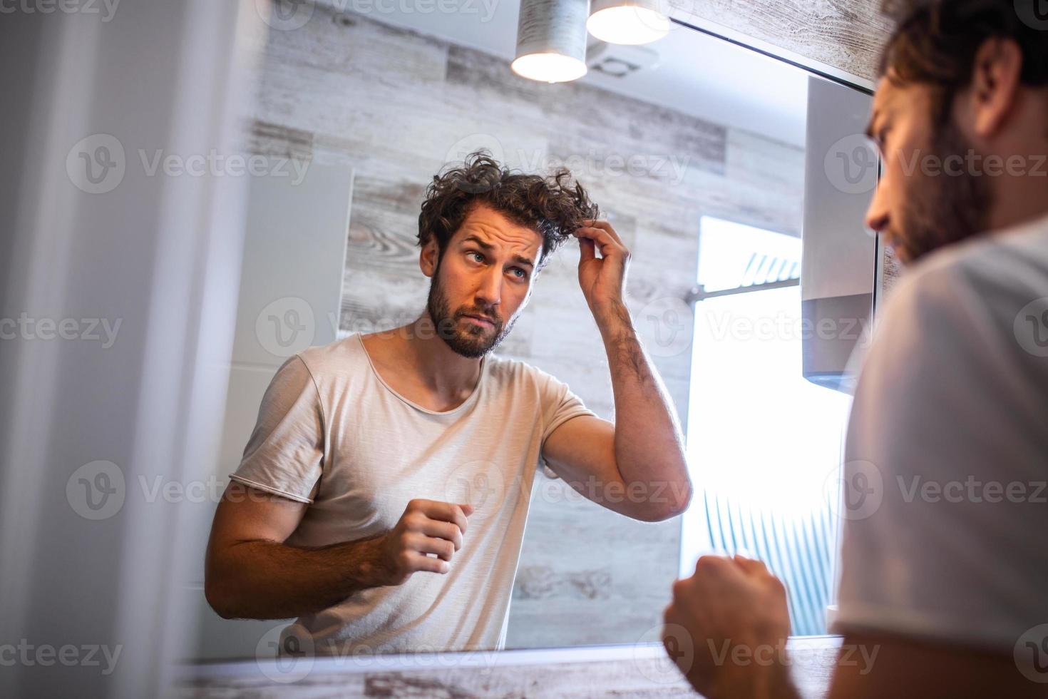 stilig ung man rörande hans hår med hand och grooming i badrum på Hem. vit metrosexual man orolig för hår förlust och ser på spegel hans avtagande hårfäste. foto