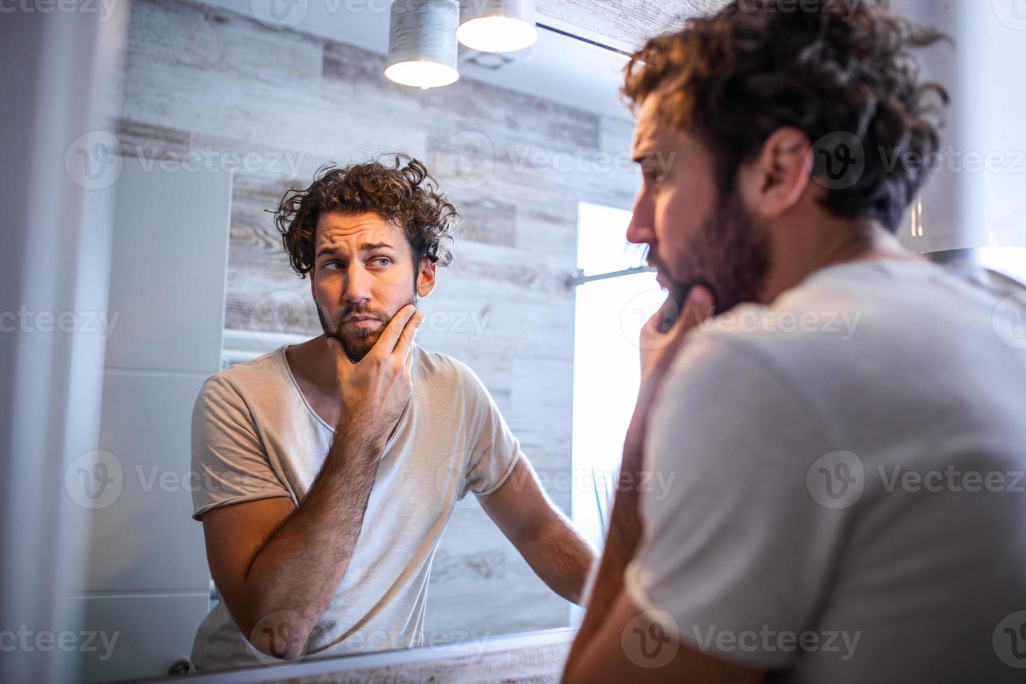 morgon- hygien, stilig man i de badrum ser i spegel. reflexion av stilig man med skägg ser på spegel och rörande ansikte i badrum grooming foto