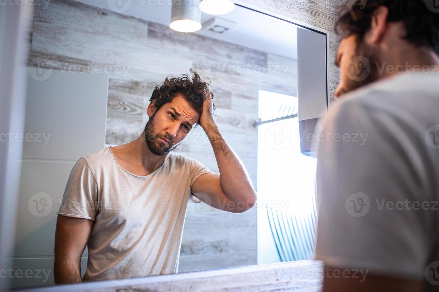 stilig ung man rörande hans hår med hand och grooming i badrum på Hem. vit metrosexual man orolig för hår förlust och ser på spegel hans avtagande hårfäste. foto