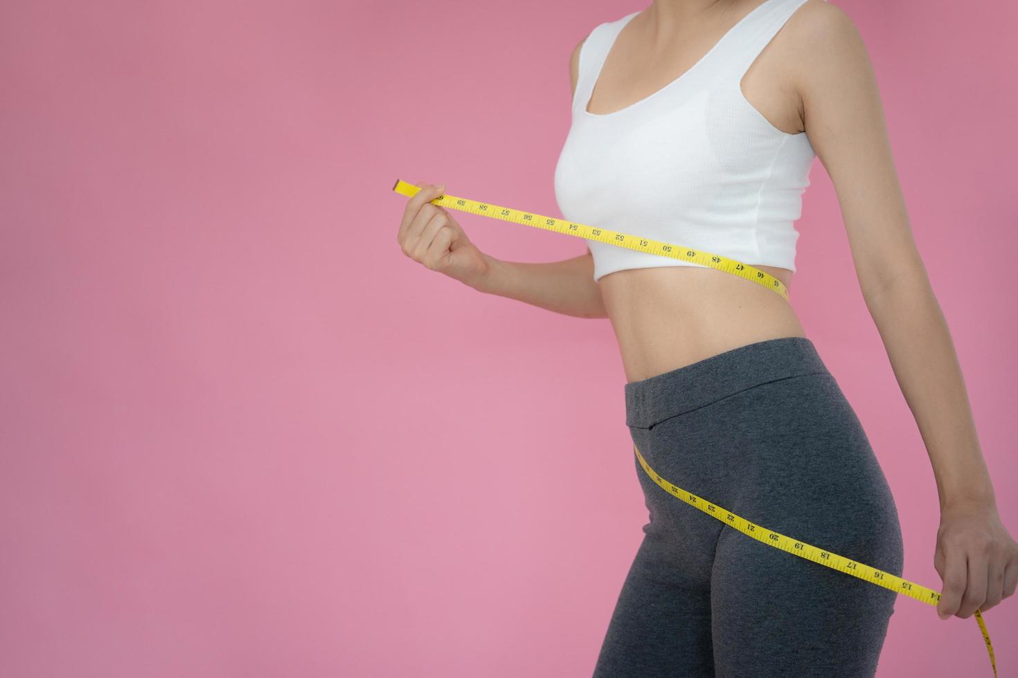 smal kvinna i sportkläder åtgärder henne midja använder sig av tejp mäta på rosa bakgrund. diet kvinna och tappa bort vikt planen foto