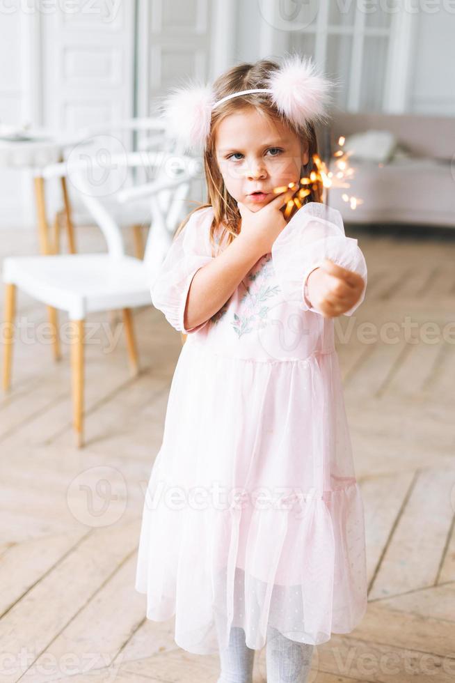 rolig söt liten flicka med lång hår i ljus rosa klänning innehav brinnande sparkler ljus i händer i ljus levande rum på de Hem. jul tid, födelsedag flicka foto