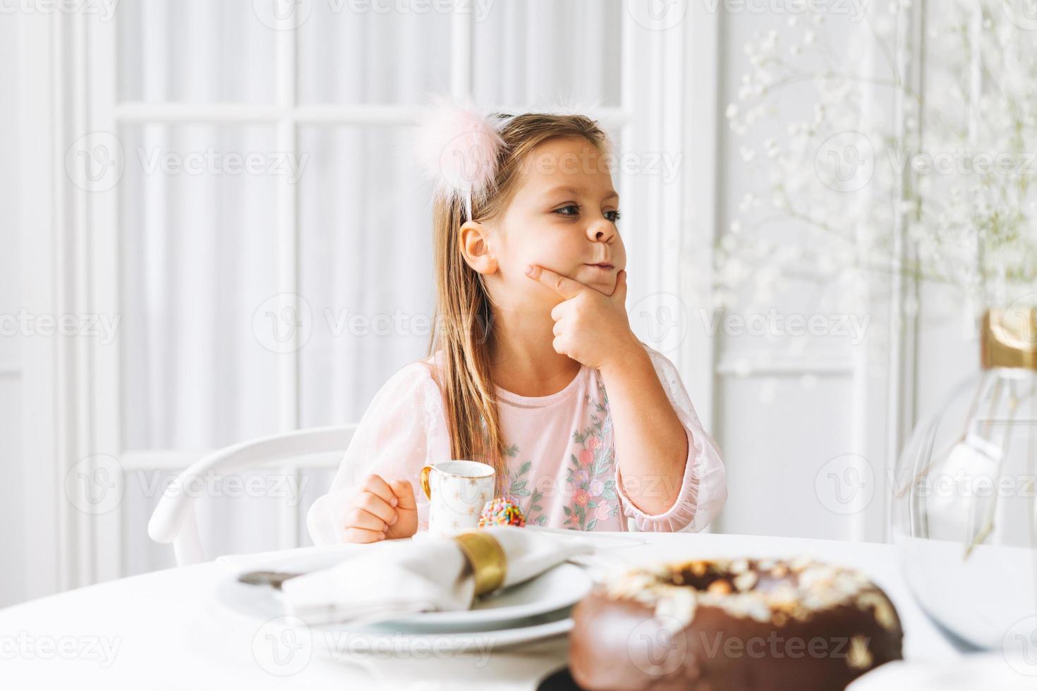 rolig söt liten flicka med lång hår i ljus rosa klänning med choklad kaka i händer på festlig tabell i ljus levande rum på Hem. jul tid, födelsedag flicka foto