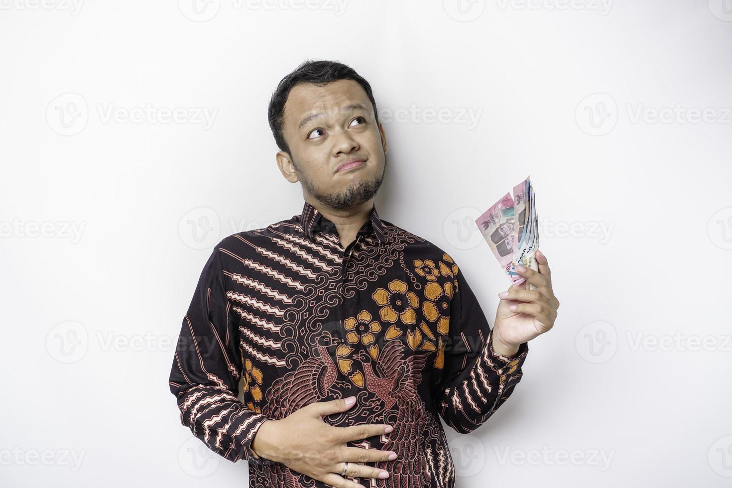 Foto av en hungrig upphetsad ung asiatisk man innehav pengar i indonesiska rupiah och undrar Vad till beställa smaskigt mat isolerat på vit Färg bakgrund