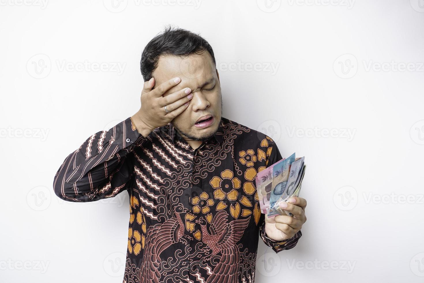 en betonade asiatisk man bär batik skjorta och innehav pengar i indonesiska rupiah isolerat förbi vit bakgrund utseende deprimerad foto