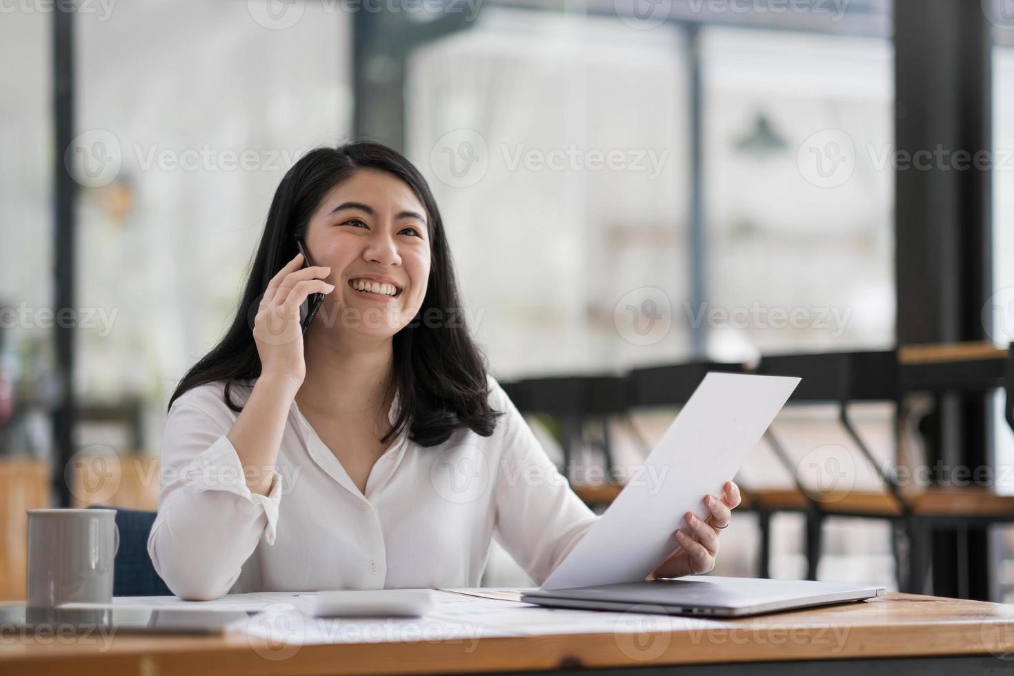 glada leende asiatisk affärskvinna som arbetar på laptop på kontoret, med smart telefon. affärskvinna sitter på hennes arbetsplats foto