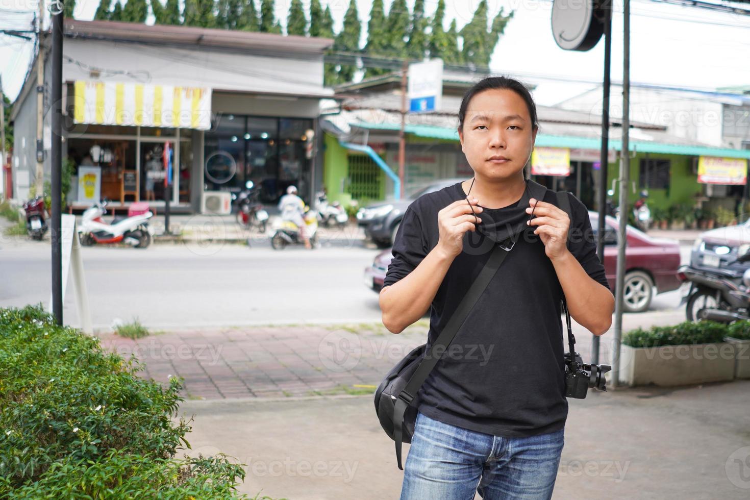 asiatisk man i svart t-shirt och jean byxor är bär svart ansikte mask i främre av blur thailand kommersiell byggnad och lantlig väg område. foto