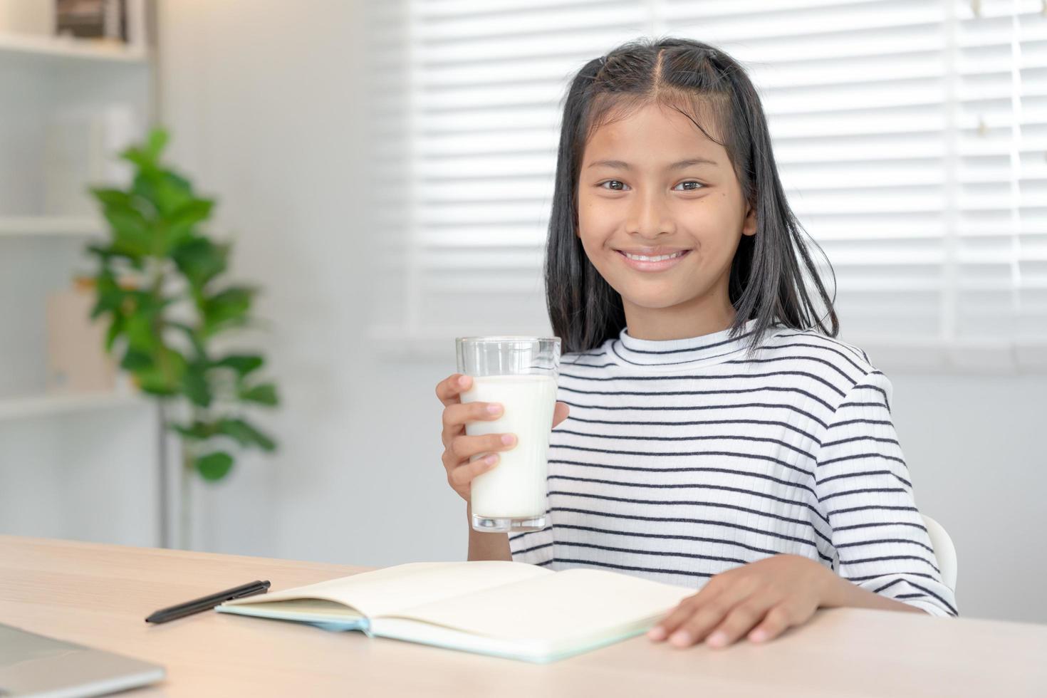 barn kvinnor dryck mjölk till uppföda de kropp och uppföda de hjärna. asiatisk ung liten flicka lära sig på Hem. flicka Lycklig dryck mjölk och läsa en bok för examen, hemskola. utbildning, vitaminer, utveckling foto