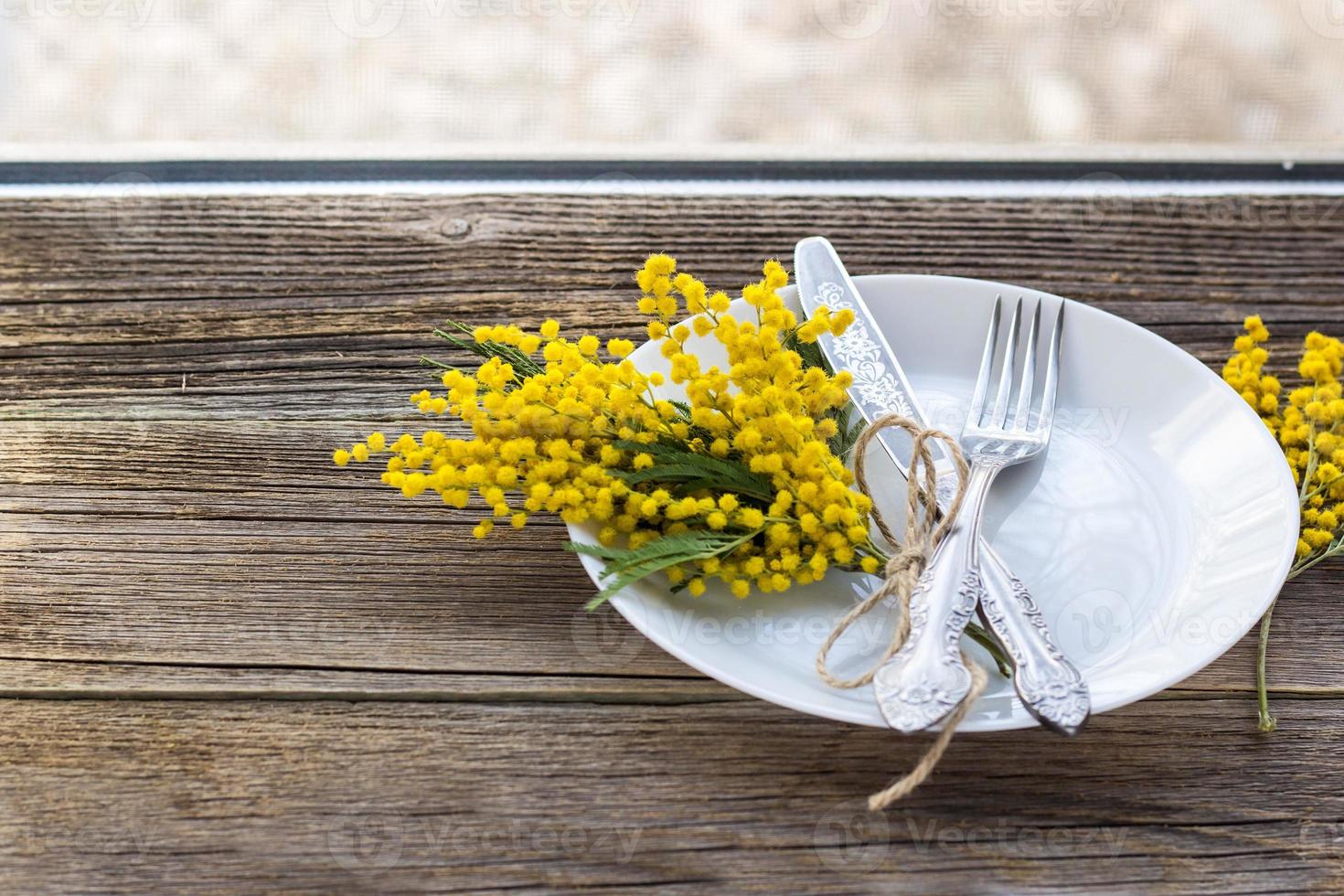 gaffel kniv med tallrik och mimosa blommor på trä- tabell på fönster bakgrund. vår påsk Semester middag. foto