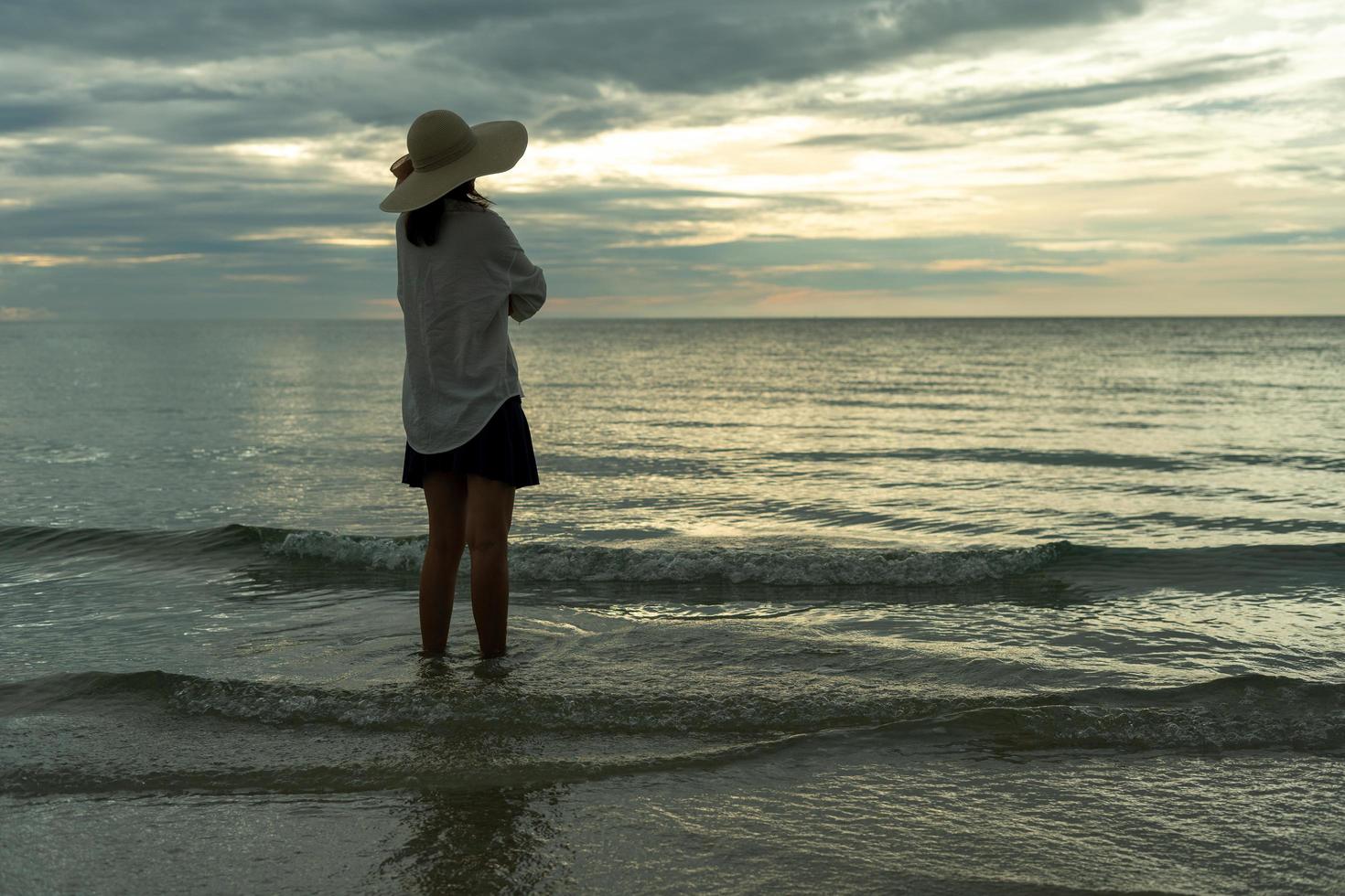 kvinna bär en hatt promenader ensam på ett tömma sandig strand på solnedgång. en ensam och deprimerad ung kvinna står på de sand av de strand på Semester. foto