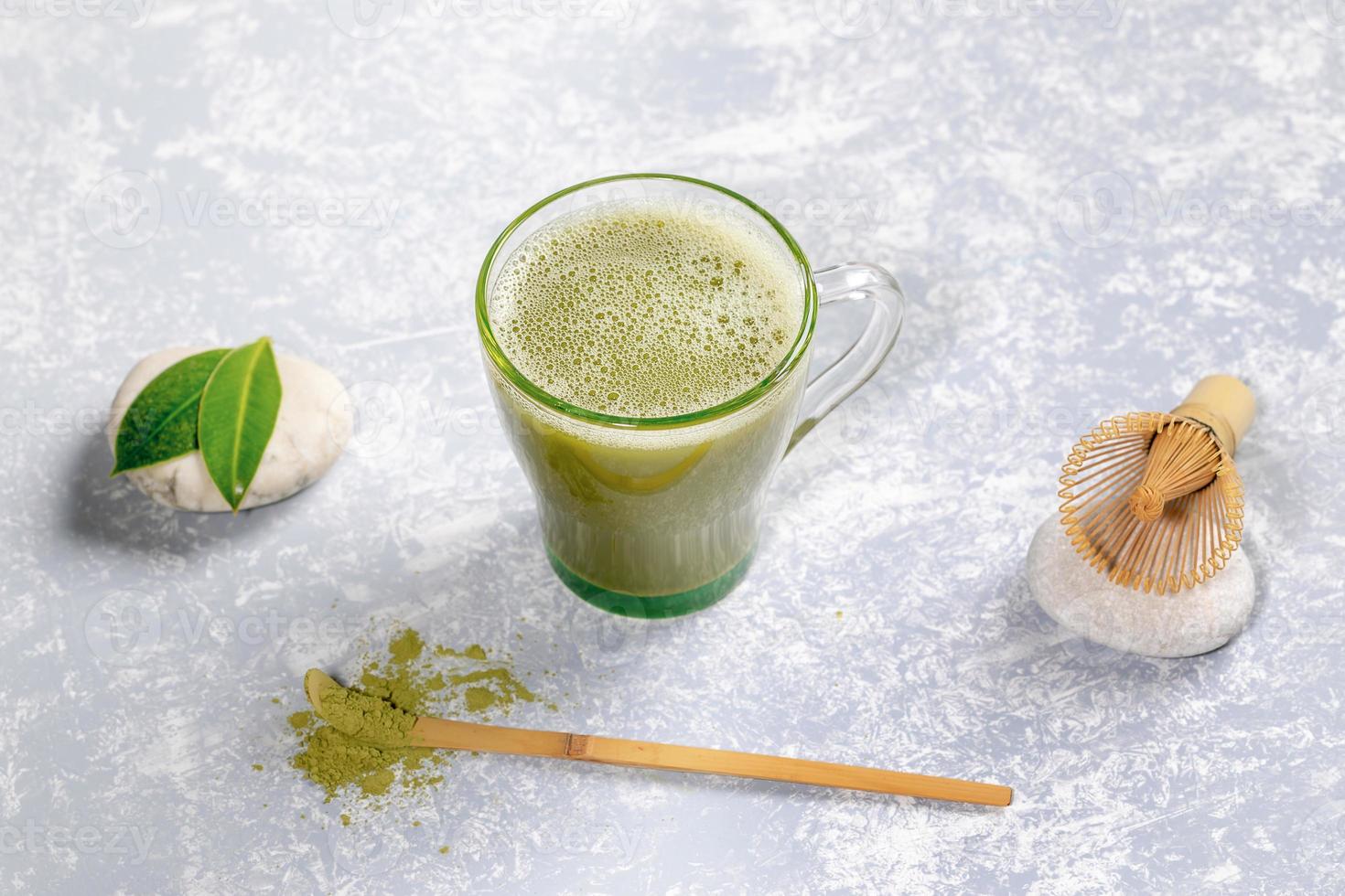 glas kopp med matcha grön te, trä- sked med pulver, bambu vispa och löv på stenar på grå texturerad bakgrund. foto