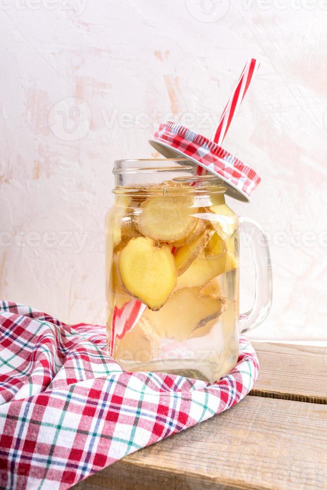 glas burk med ingefära vatten med dricka sugrör och röd tartan keps på röd rutig trasa på trä- plankor på ljus bakgrund. foto