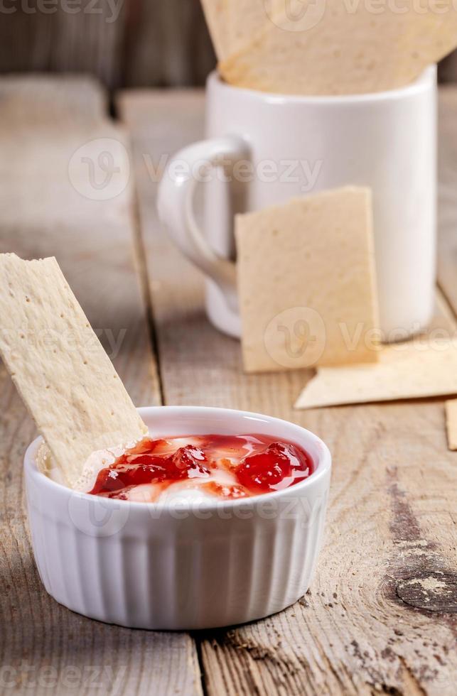 närbild keramisk skål med sur grädde och jordgubb sylt med vete hårt bröd på gammal trä- tabell. foto