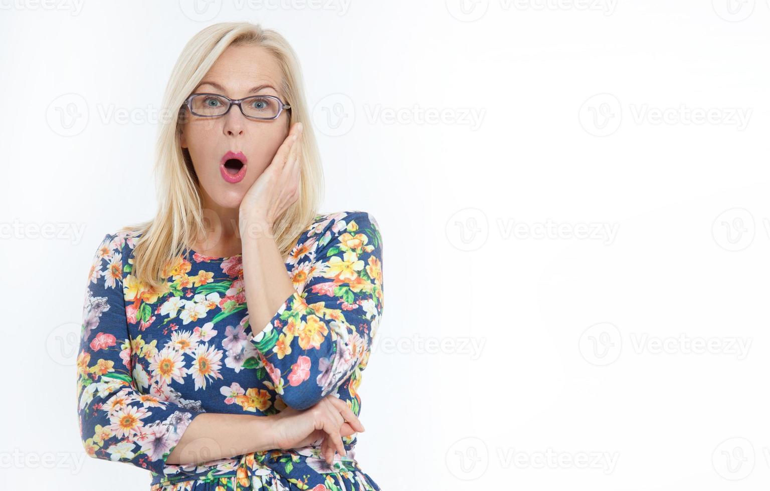 Lycklig kvinna med glasögon och överraskad ropar innehar kinder förbi hand. skön kvinna med blond hår pekande till se. presenter din produkt. uttrycksfull ansiktsbehandling uttryck medelålders kvinna. foto